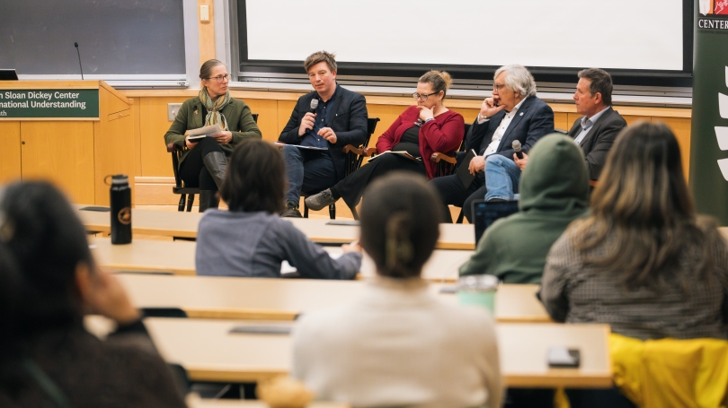 The camera looks between people seated in a lecture theater, and focusses on three individuals in seats. One of them is holding a microphone and speaking.