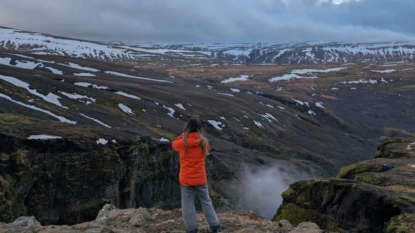 The camera is pointed at the back of a student in a bright coat, as they look out across a mountainous landscape
