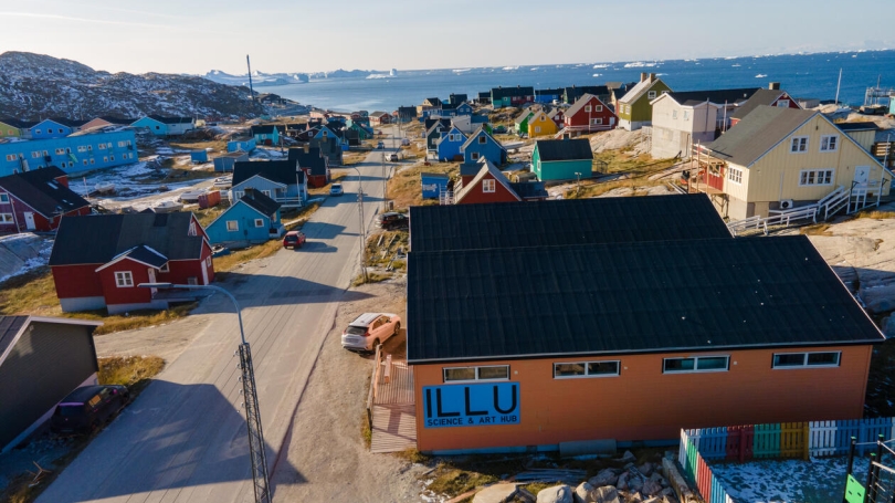 A drone-level photo that looks out on colourful houses in Illulisat, Greenland. The building in the foreground is the ILLU Science and Art Hub. Visible in the distance is the ocean.