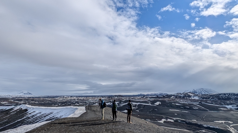Three students wearing coats standing on the rim of a dead volcano crater, with mountains in the background.jpg
